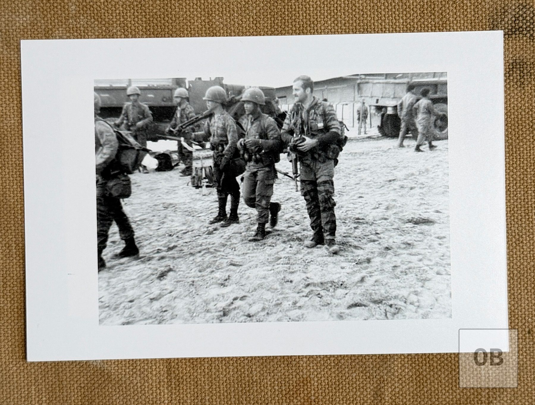 Ulricson in tigerstripe camouflage carrying his rifle and gear across a sandy compound beside helmeted Montagnard soldiers, with trucks behind them.
