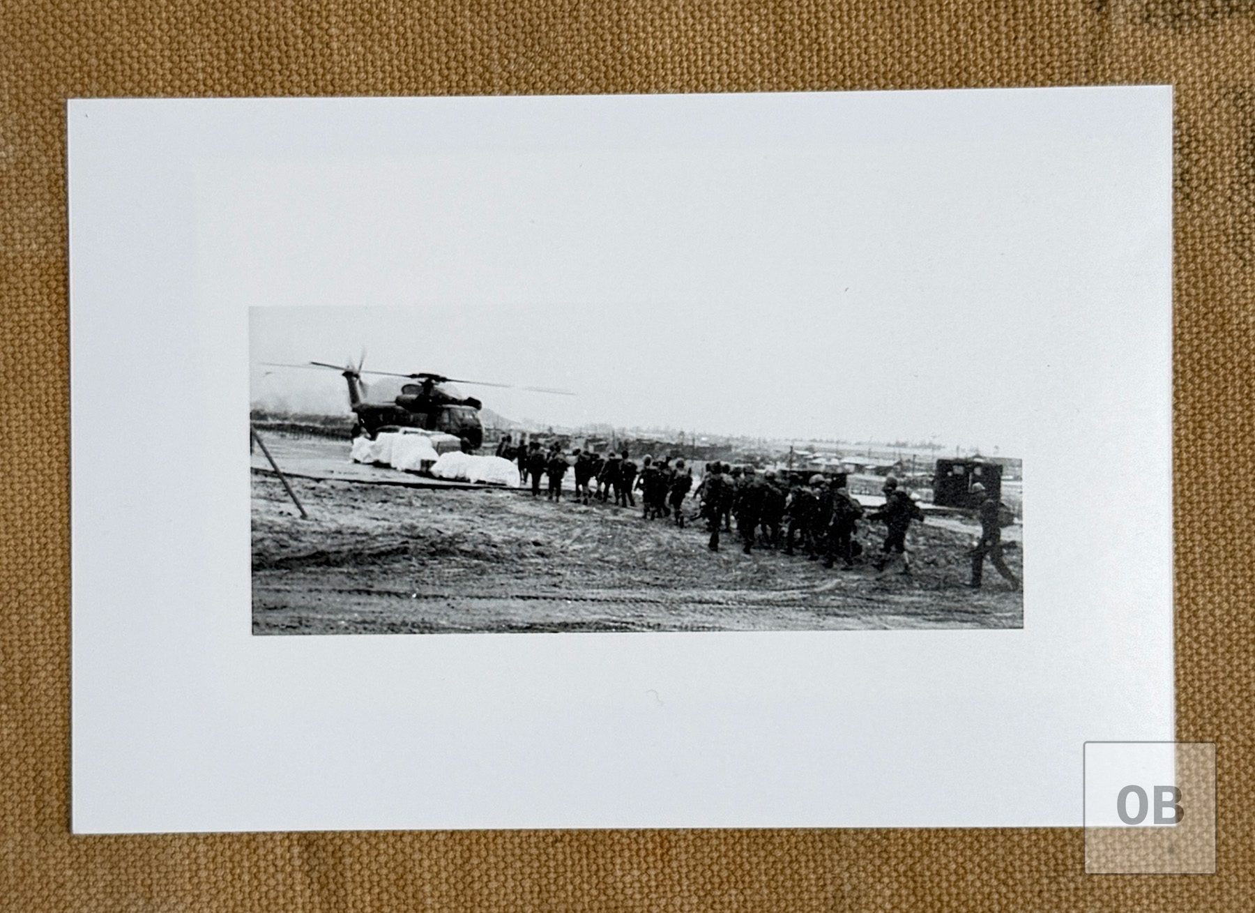 A column of soldiers walking across an airfield toward a large helicopter, with covered cargo pallets beside it.