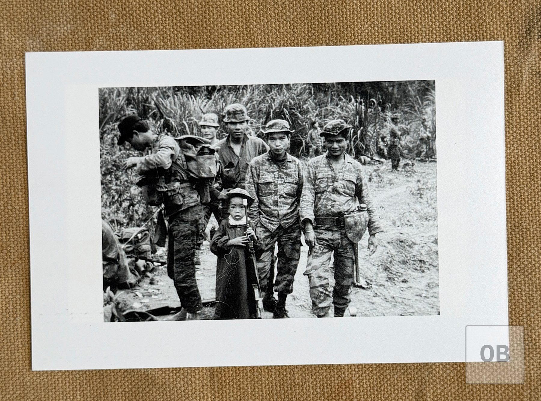 A small Montagnard boy in an oversized jacket and bush hat holding an M16 rifle, standing with Montagnard soldiers in tigerstripe camouflage.