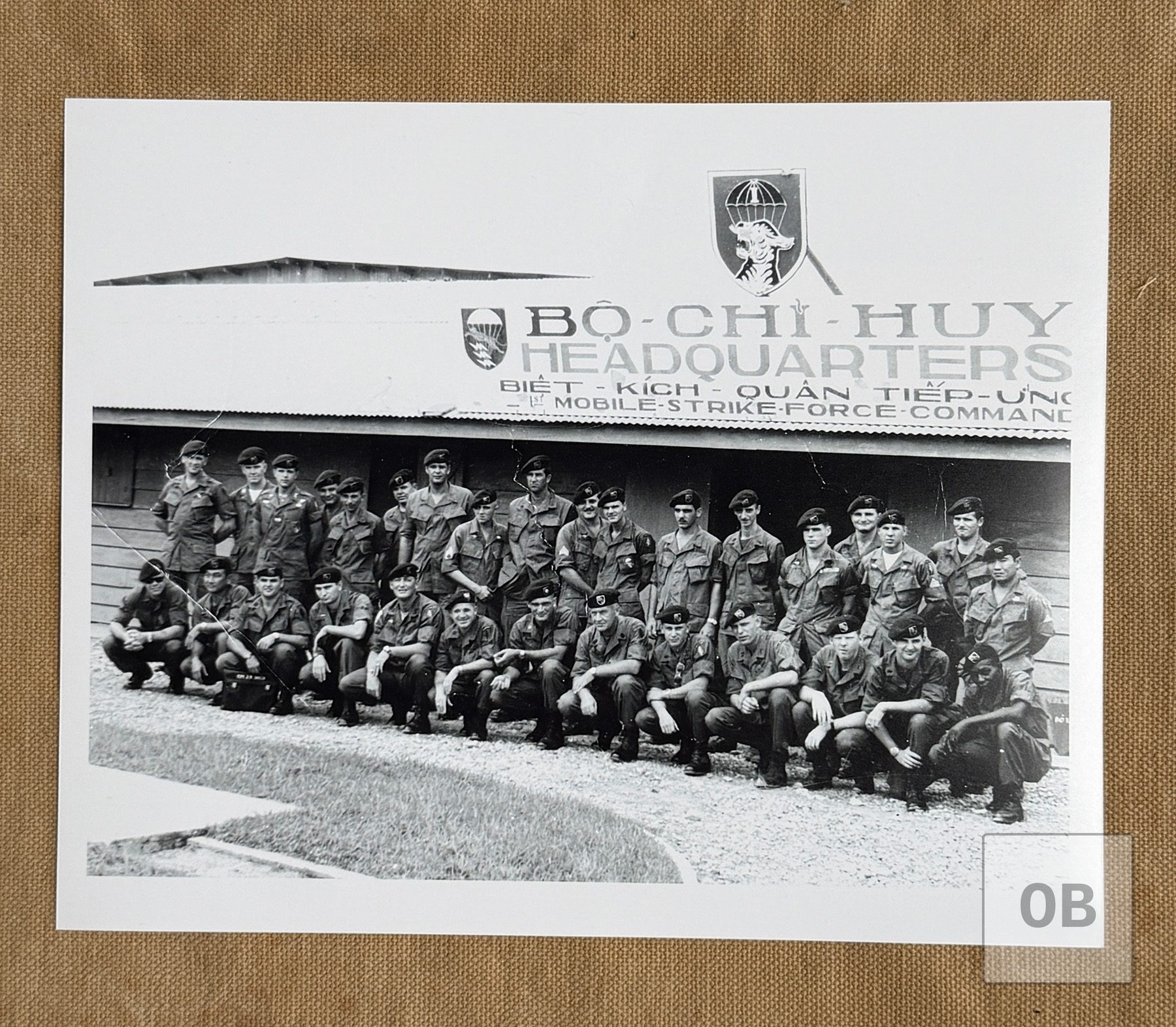 Group photograph of Special Forces soldiers in berets, standing and kneeling in two rows before a building signed Bo Chi Huy, Headquarters, Biet Kich Quan Tiep Ung, 1st Mobile Strike Force Command.