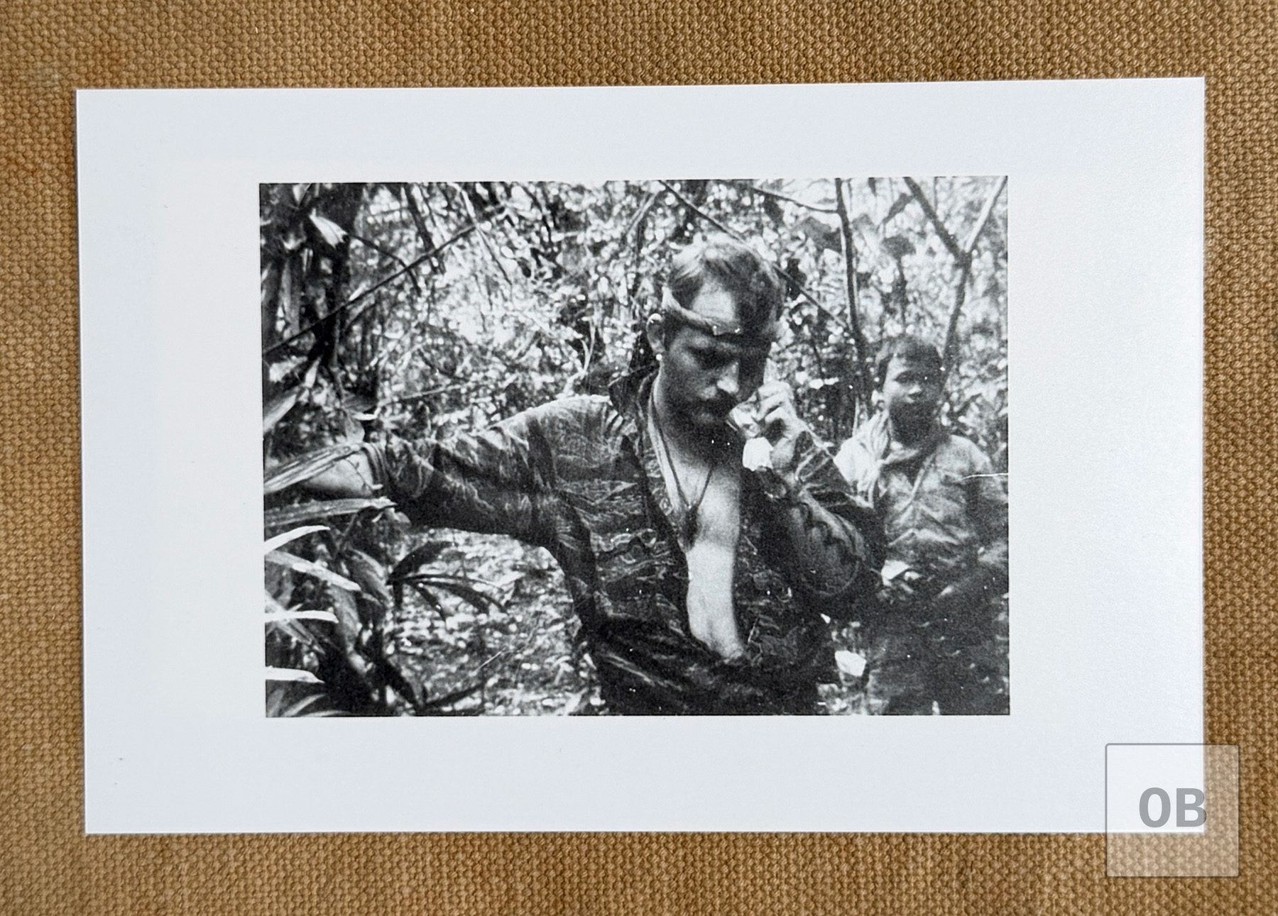 Ulricson in an open tigerstripe shirt and headband speaking into a radio handset in the jungle, with his young Montagnard radio operator behind him.