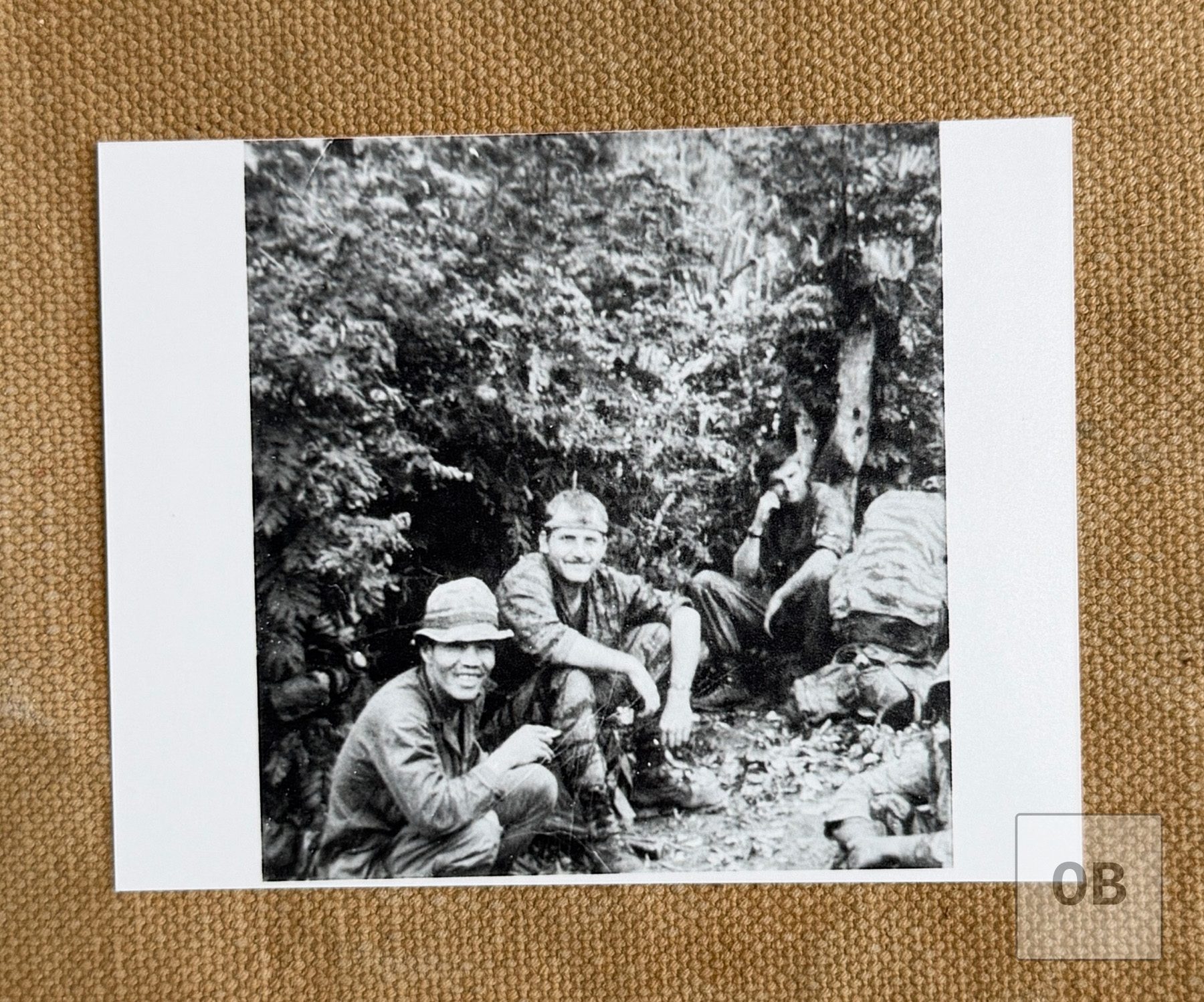 Ulricson in tigerstripe camouflage seated in the jungle beside a smiling Montagnard soldier in a bush hat, with other soldiers resting nearby.