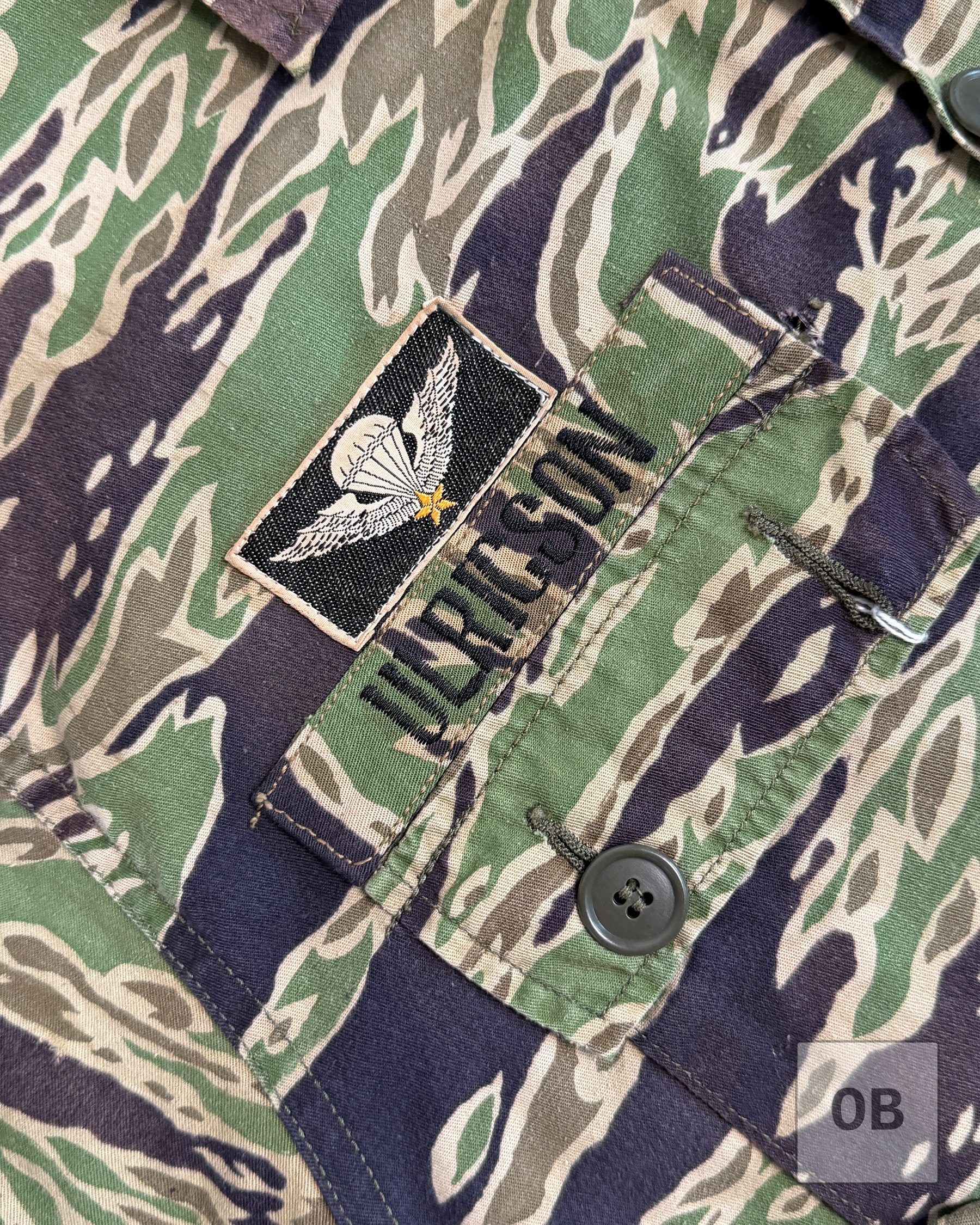 Close-up of the machine-sewn ULRICSON name tape in lightweight tigerstripe material, with South Vietnamese silk woven jump wings on a black background above it.